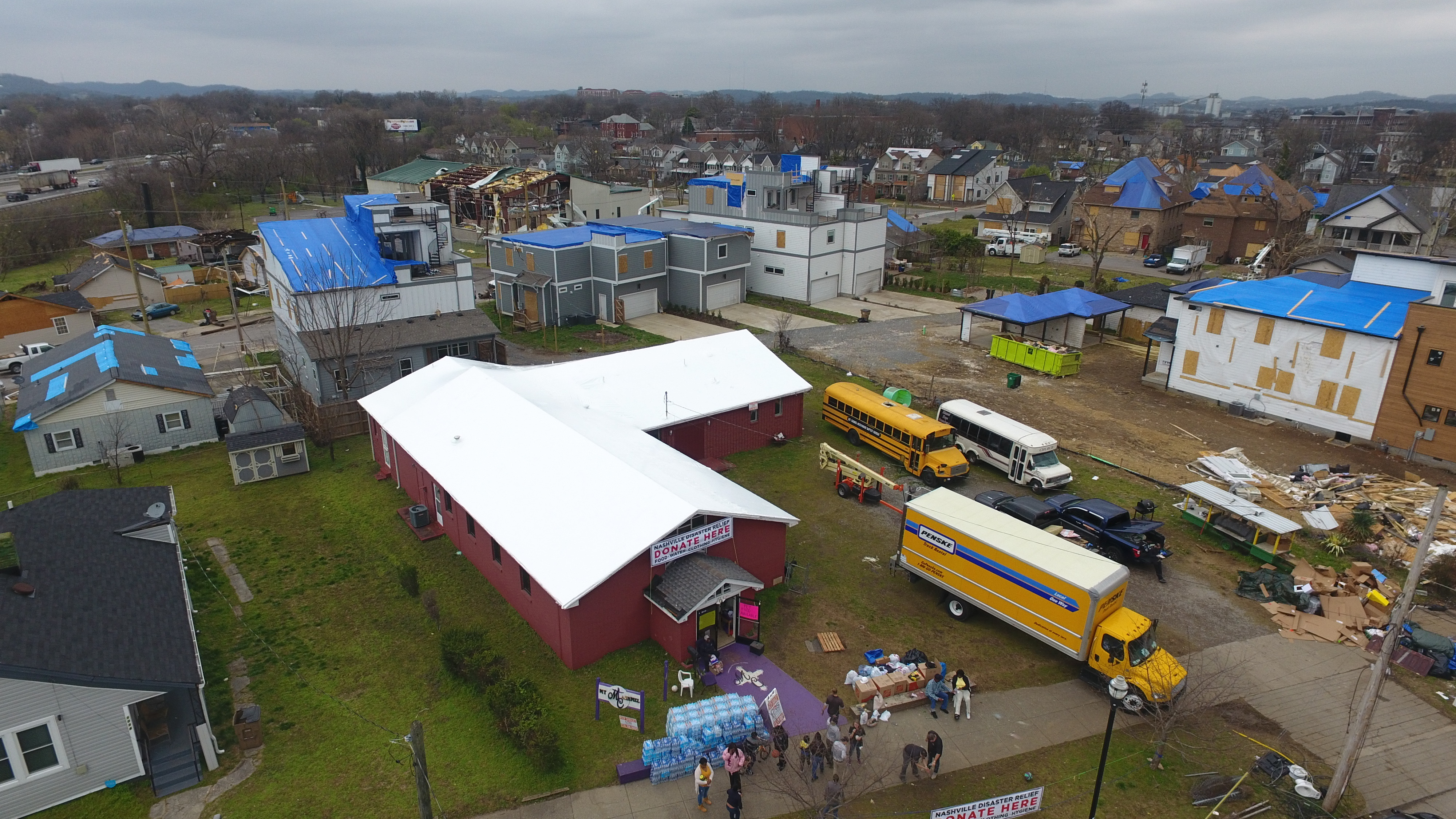 Aerial view of a storm-hit neighborhood, one building sealed under white RoofWrap surrounded by homes still under blue tarps